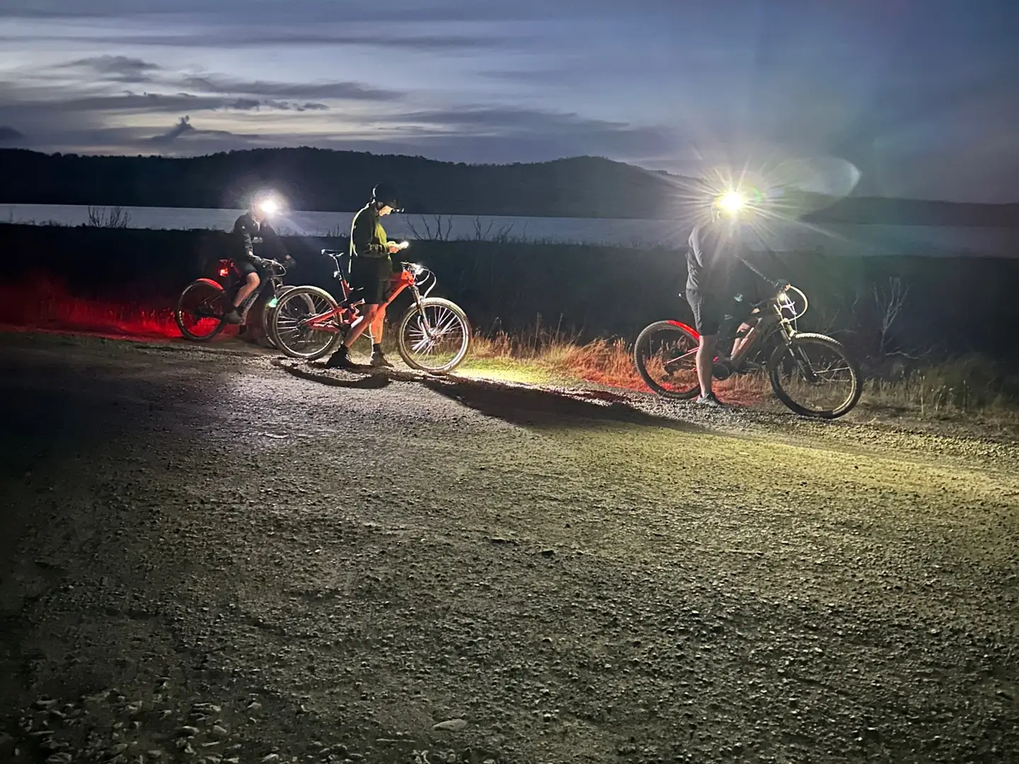Three riders stopped with their bikes and lights beside water at dusk