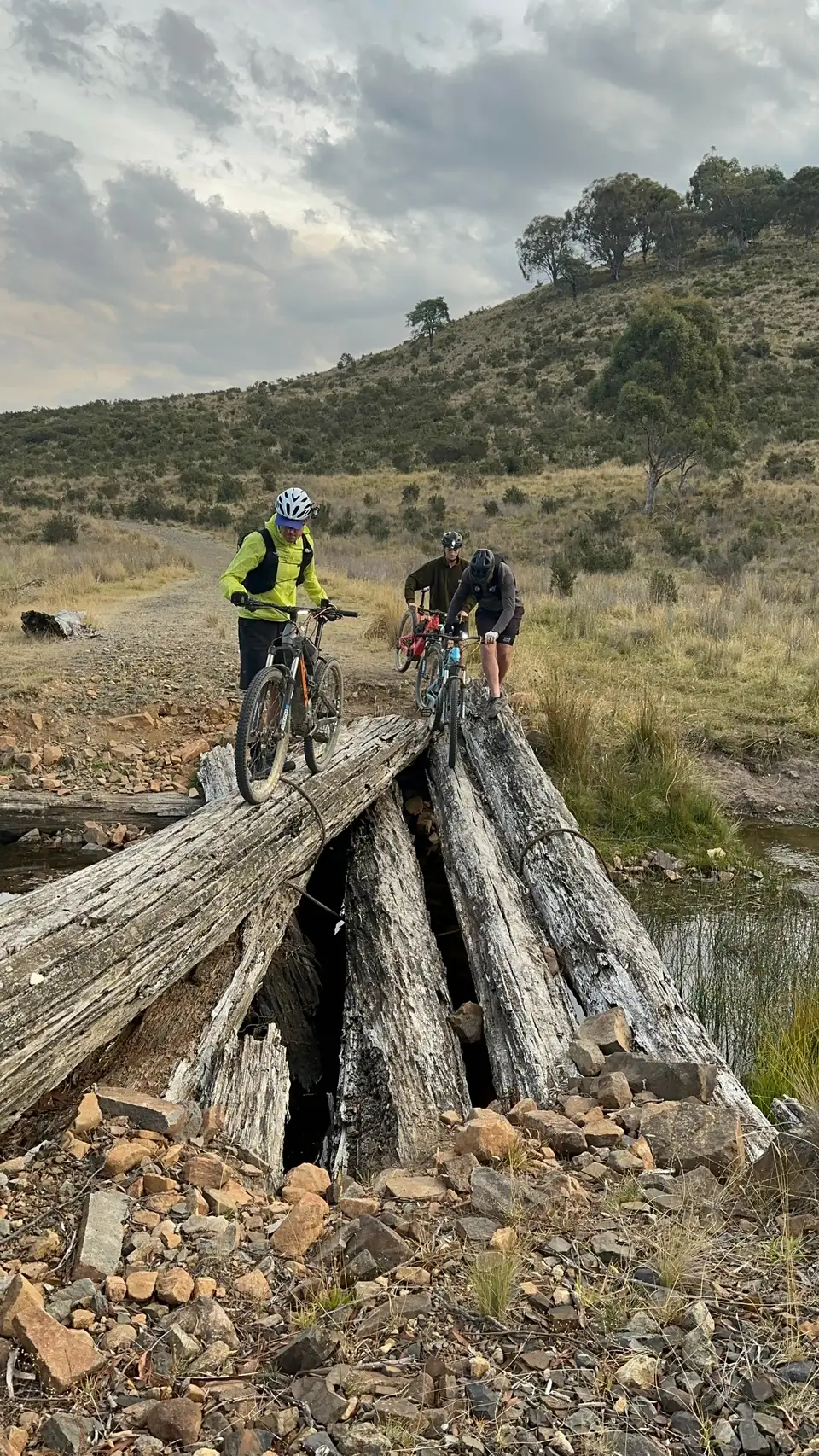 Helmeted riders walking their bikes across a weathered log bridge